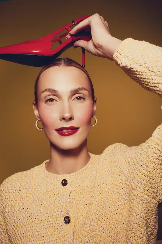 Young woman with red lipstick and hoop earrings holds red high heel shoe above her head. She wears a beige knitted sweater against a warm brown background. Fashion and style concept.