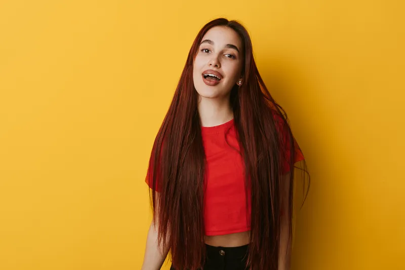 Young woman with long red hair wearing red crop top smiling and posing in front of yellow background. Casual style, confident expression and natural look for portrait photography.