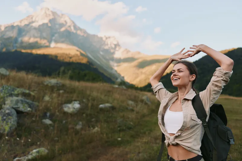 A woman outdoors, hiking near majestic mountains in daylight, wearing a backpack and casual attire, enjoying freedom, joy, and an active lifestyle in nature.