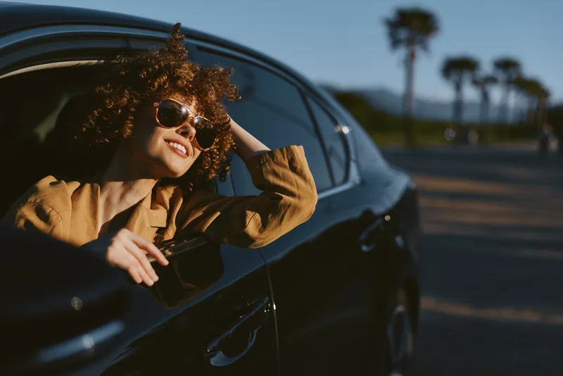 A young woman leans out of a car window, wearing a brown jacket and sunglasses, basking in warm sunlight while palm trees blur by in the background.