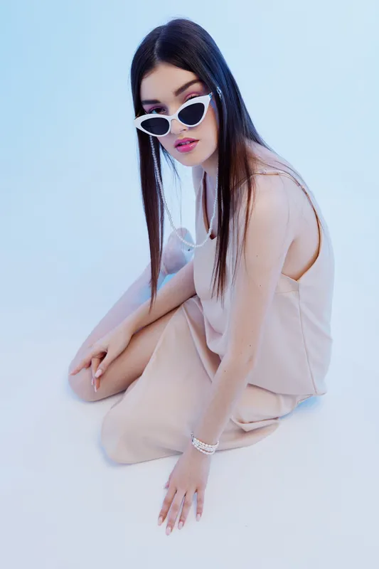 Young woman in beige sleeveless dress and white sunglasses sitting on the floor against a blue background. Concept of beauty, fashion, style, and modern trends in studio lighting.