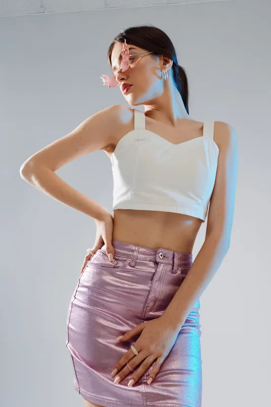 Young woman in a white crop top and shiny pink skirt poses with hand on hip and flower near face. Studio shot with soft lighting. Concept of woman beauty fashion and elegance.