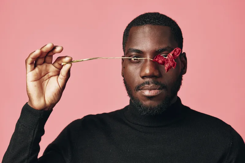 African man with dark skin holding red flower over eye in creative conceptual visual style on pink background. Portrait of confident black male with artistic expression and fashion look.