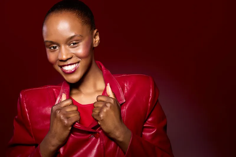 Confident black woman with dark skin wearing a red leather jacket smiles brightly on a red background. Portrait of a stylish African female model expressing happiness and self-assurance.