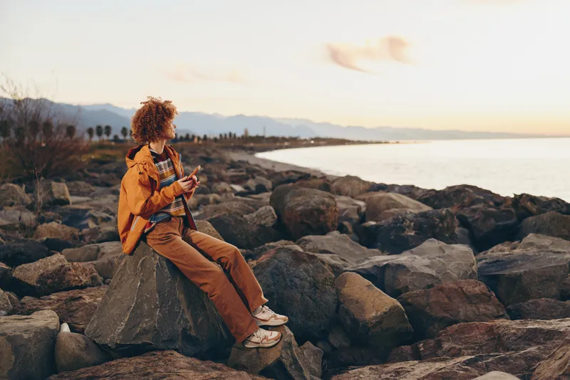 Woman lifestyle on seaside rocks at sunset, coastal ocean waves and warm sky. She sits on the shoreline in an orange jacket, travel mood, outdoor scene with horizon and calm water.