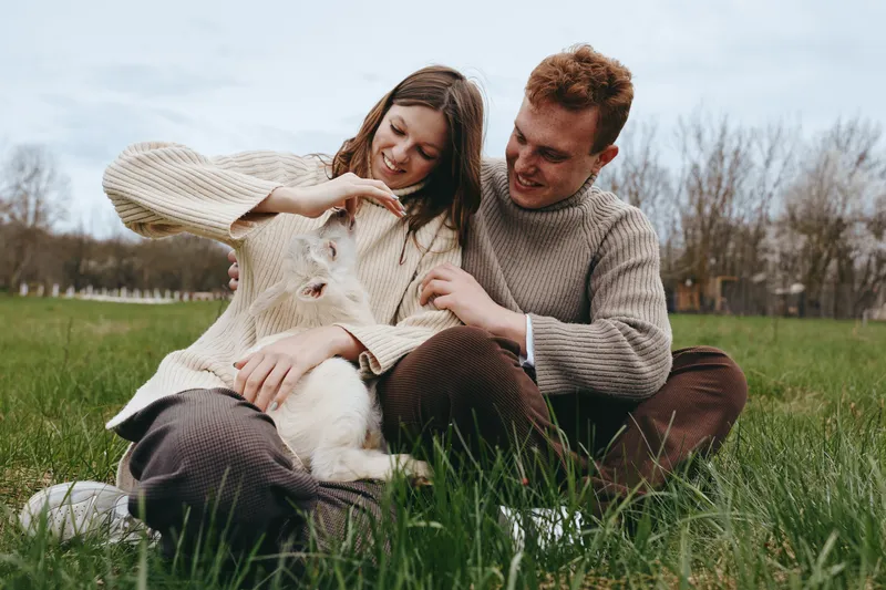 A couple sits on the grass in a sunny park, stroking a friendly dog as they share a light, affectionate moment. A lifestyle scene capturing companionship, outdoor activity, and relaxed, joyful