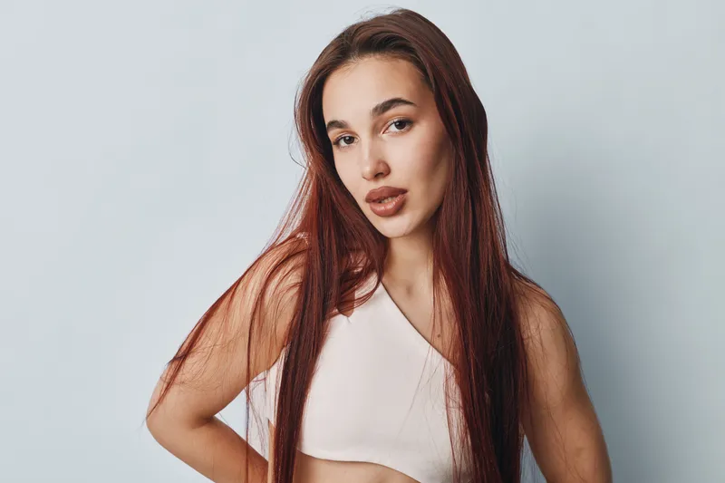 Woman with long red hair and natural makeup posing in white top against plain background. Portrait of confident female with hand on hip and relaxed expression in studio lighting.