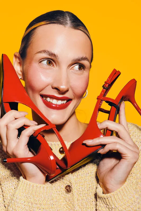 Smiling woman with red lipstick holds red high heel shoes near face on yellow background. Concept of fashion, style, beauty, and shopping enthusiasm.