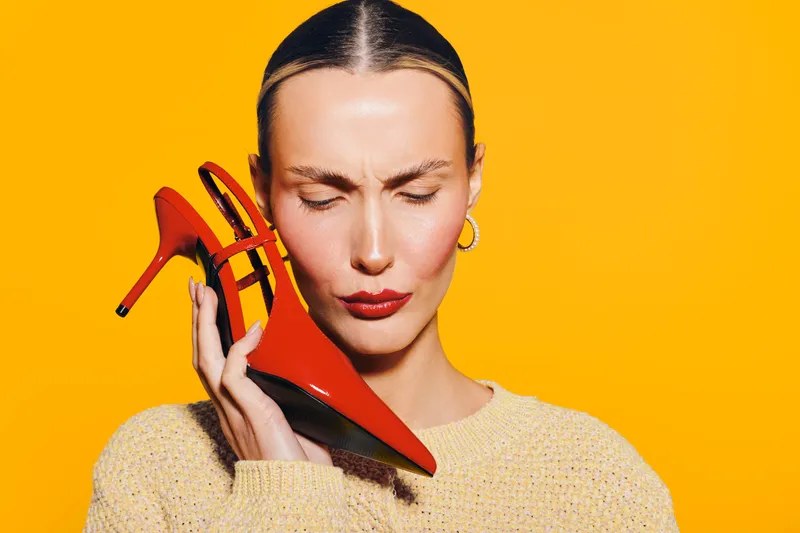 Woman with closed eyes holds red high heel shoe near face on bright yellow background. Concept of fashion, style, and bold color contrast in modern portrait photography.