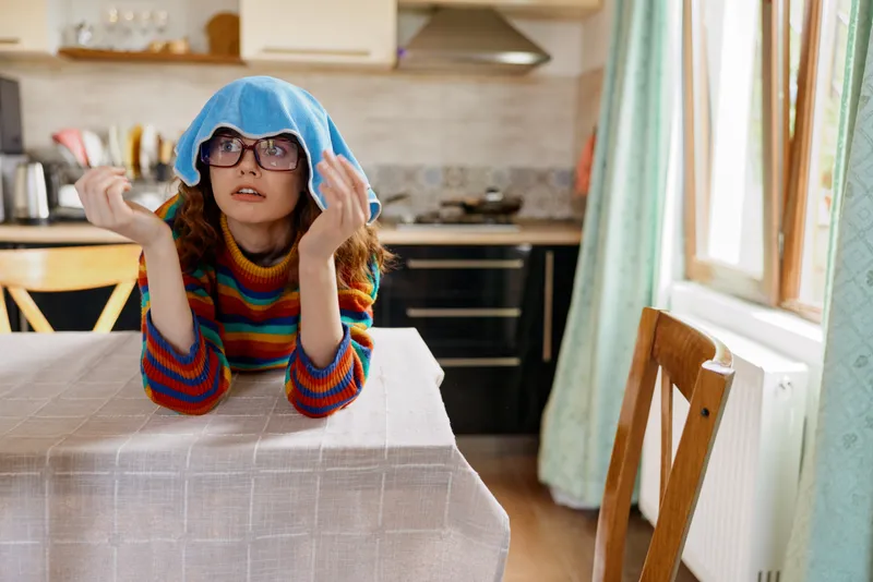Cheerful girl indoors at a home kitchen table, wearing a rainbow sweater and blue hood, playful pose, light streaming in, candid lifestyle moment, warm atmosphere