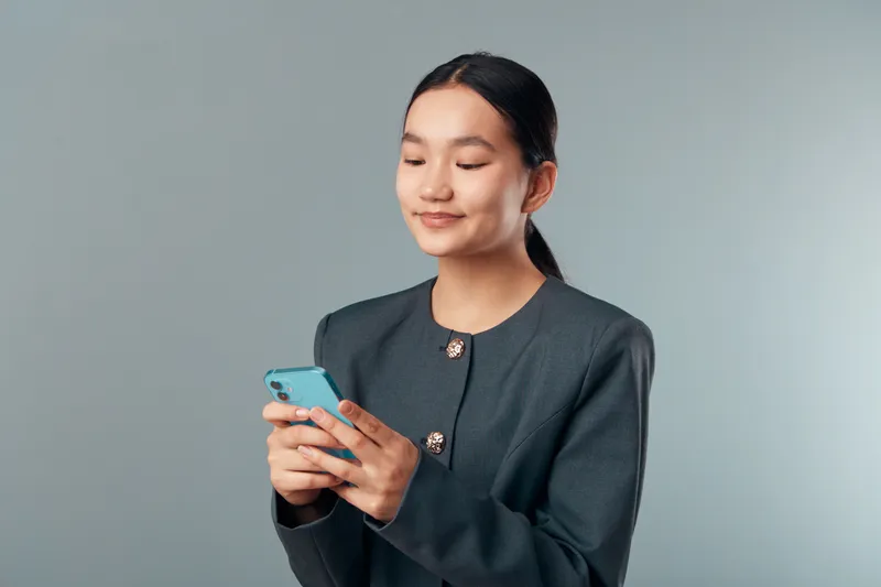 Young woman in formal blazer smiling and using smartphone with both hands standing against plain background. Technology, communication and mobile phone for digital interaction and social media.
