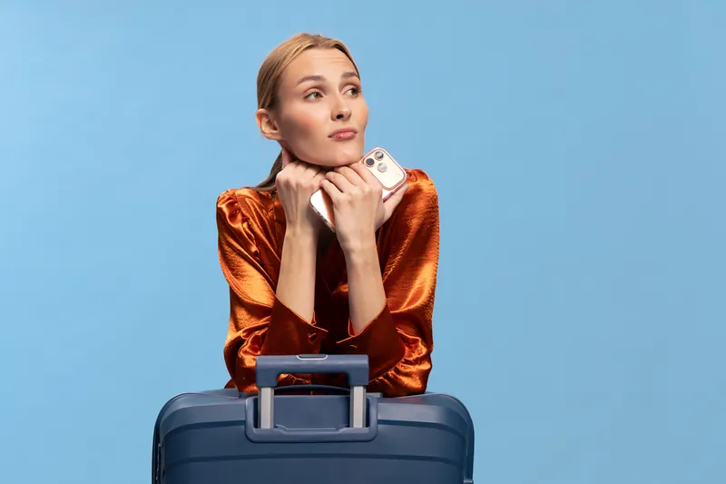 Young woman in orange blouse leans on blue suitcase holding smartphone, looking away thoughtfully on blue background. Concept of travel, waiting, planning, and modern lifestyle.