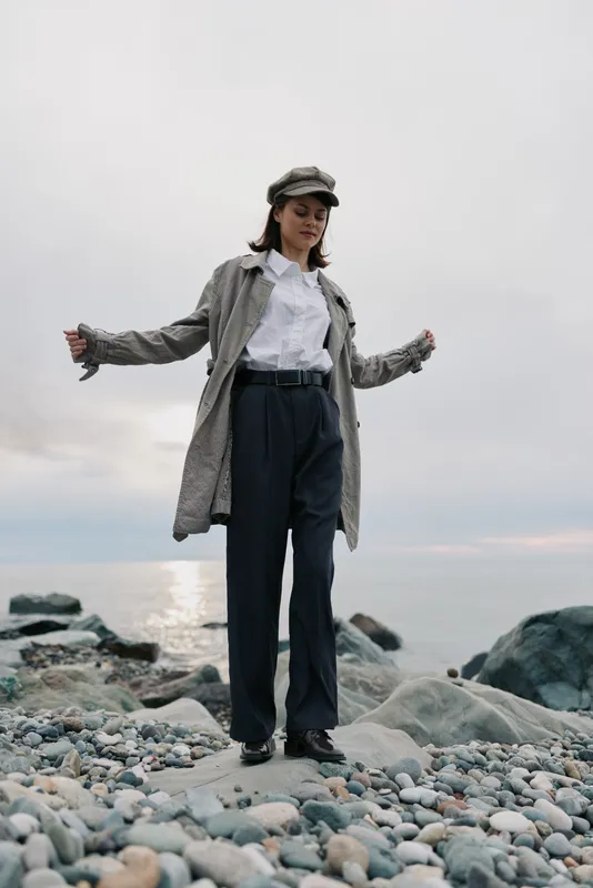 Young woman in grey coat and cap stands on rocky beach near calm sea at overcast sunset. She wears white shirt and dark pants, posing with arms outstretched for fashion and nature concept.