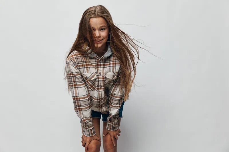 Girl with long hair wearing plaid shirt and denim shorts standing and smiling on white background. Youthful fashion casual style with natural expression and windblown hair.