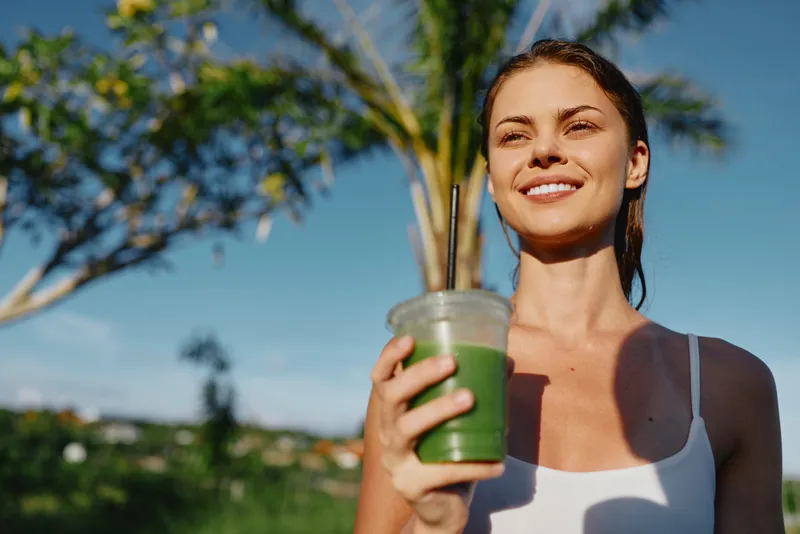 Young woman wearing a white tank top holds a green smoothie outdoors, smiling and enjoying a sunny day with palm trees and a bright blue sky behind her.