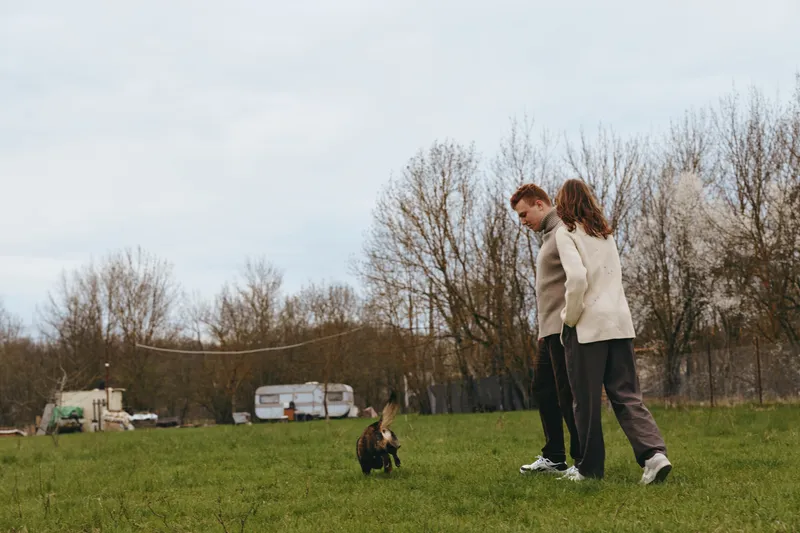 Lifestyle scene with two people walking in a grassy field, observing a small goat kid nearby. Natural daylight and relaxed mood convey bonding, outdoor life, and gentle countryside moments.