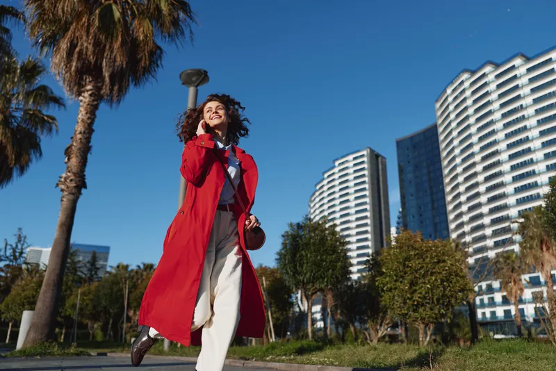 Young woman with curly hair wearing a red coat walks confidently in an urban park with modern buildings and palm trees in the background.