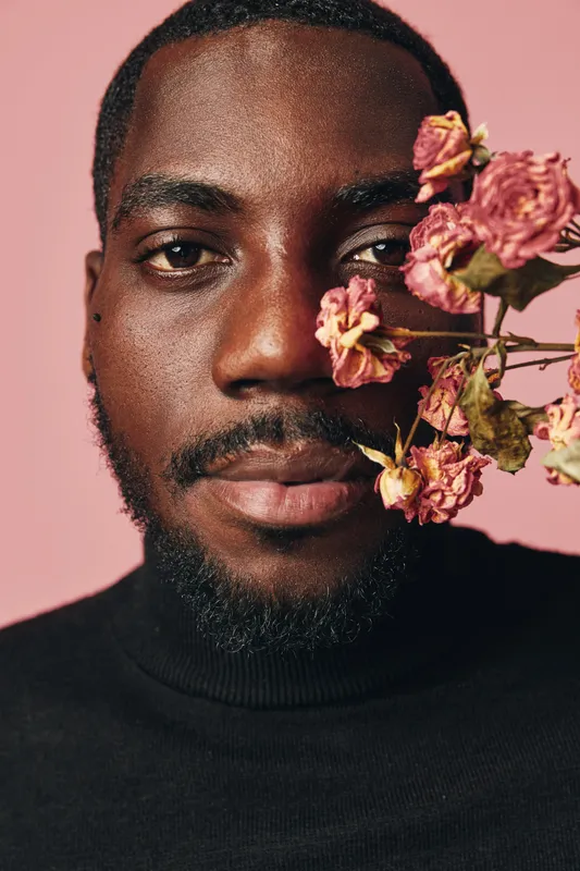 African man portrait with dried pink flowers on face in creative conceptual visual style. Closeup of black male model with beard wearing black turtleneck on pink background.