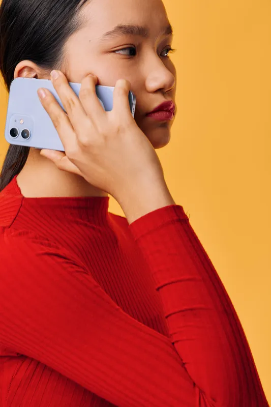 Young woman in red sweater holding smartphone to ear with serious expression on yellow background. Portrait of female using mobile phone for communication and conversation indoors.