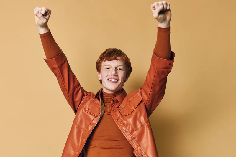 Happy young man with red hair wearing brown leather jacket and turtleneck raising arms in celebration on beige background, smiling and expressing joy and success.