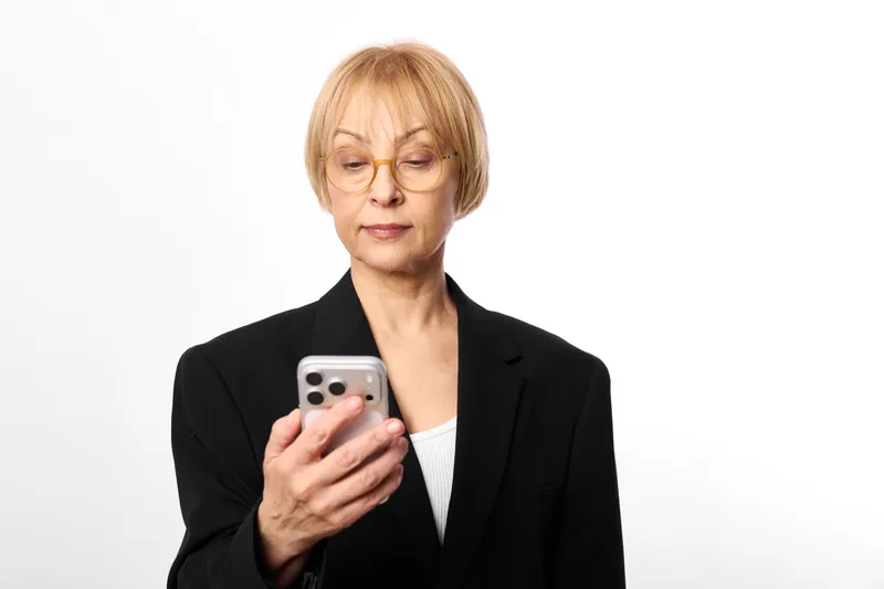 Senior business woman holding smartphone with focused expression, mobile communication, head and shoulders portrait on clean white background, minimal office look, copy space calm