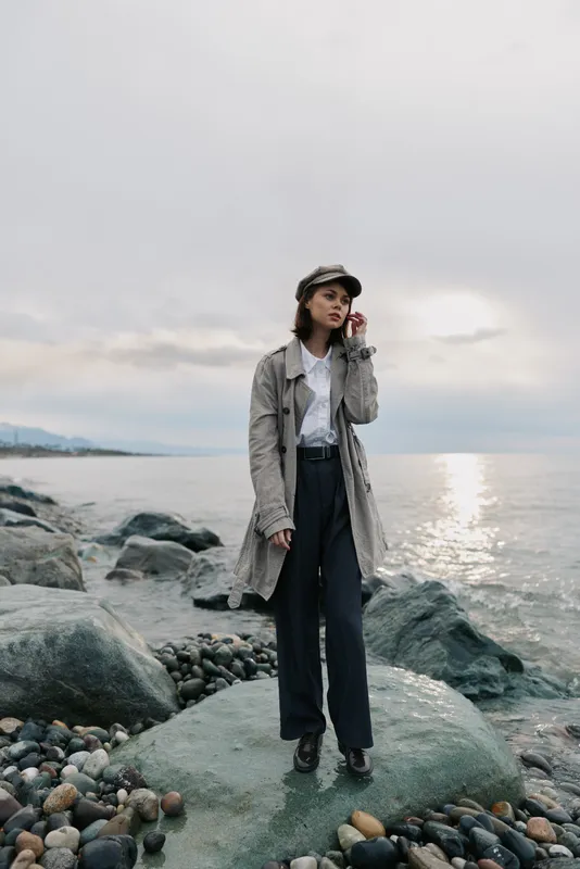 Young woman in a long coat and hat stands on rocky shore near calm sea during cloudy day. Fashionable outfit and casual pose create a moody coastal lifestyle concept.