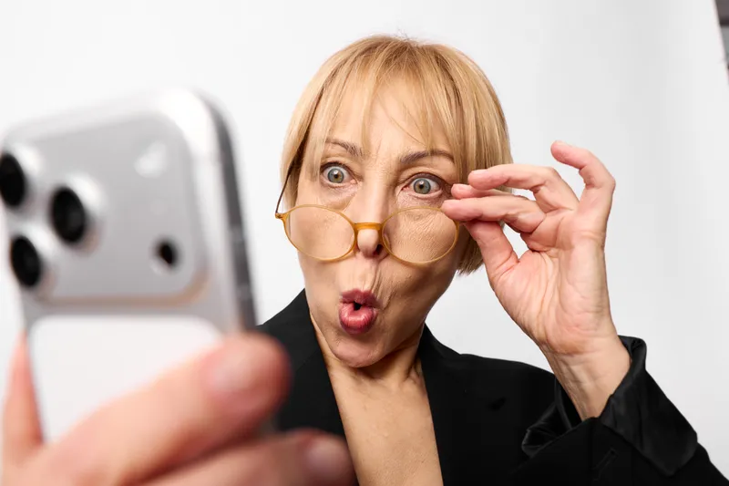 Surprised woman applying under-eye patches with her hands, close-up portrait in a bright studio. Skincare, beauty routine and self-care concept, smartphone blurred nearby.