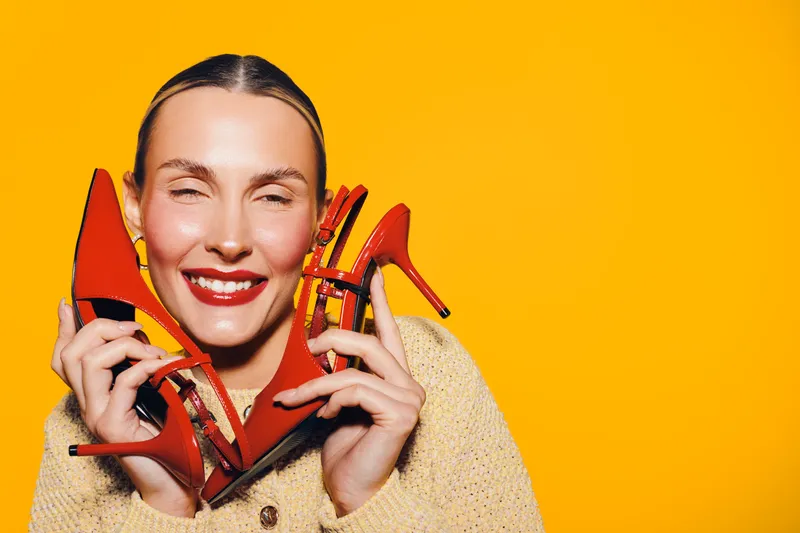 Smiling woman with red lipstick holds red high heel shoes near face on bright yellow background. Concept of fashion, style, beauty, and happiness with vibrant colors and joyful mood.