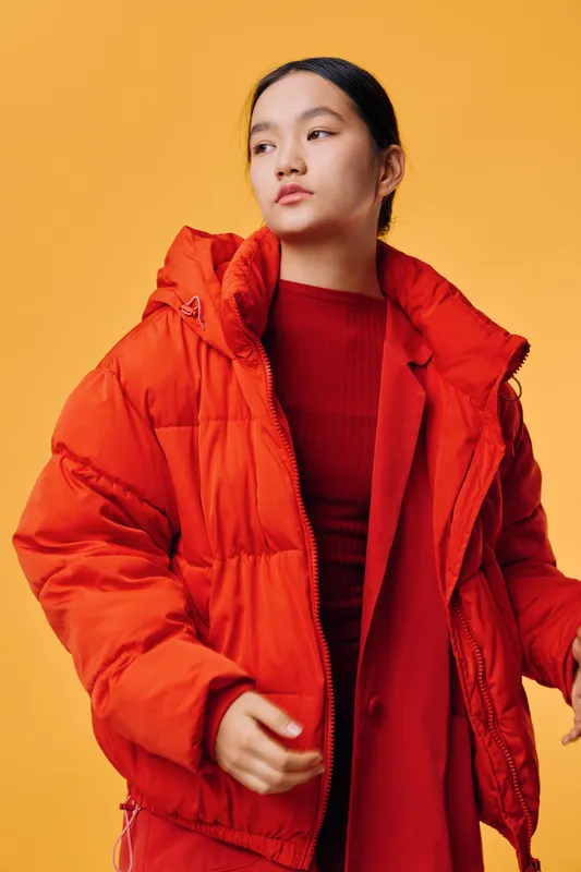 Young woman wearing oversized orange winter jacket and red outfit posing against orange background. Female model with long hair, warm clothing, fashion style and confident expression.