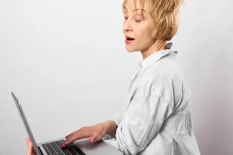 Woman with laptop in office, side profile, stress and fatigue, typing at desk in white blouse, remote work, tired mood, concentration, focus well during online tasks