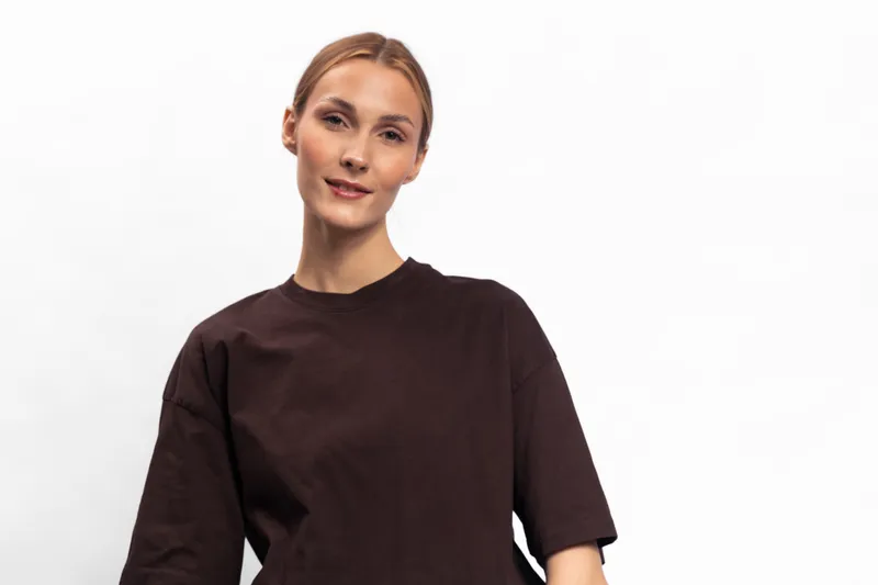 Confident young woman with tied hair wearing a loose dark brown shirt stands against white background. Studio portrait with natural makeup and relaxed posture. Concept for fashion and lifestyle.