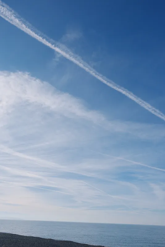 Clear blue sky above calm sea with distant coastline and white clouds, long contrails crossing the horizon, serene seascape of openness and quiet weather.