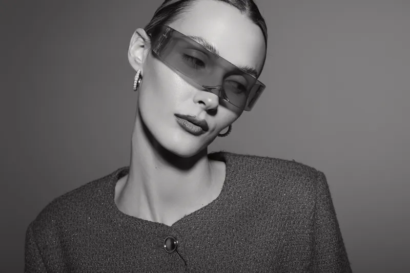 Caucasian woman with light skin wearing modern sunglasses and hoop earrings poses with closed eyes in studio. Concept of fashion, style, elegance, and minimalism in black and white.