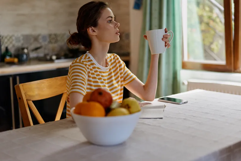 Woman enjoying a home breakfast scene, sipping from a mug, fruit bowl on the table, natural daylight from the window, casual striped shirt, peaceful morning mood, warm kitchen atmosphere, soft