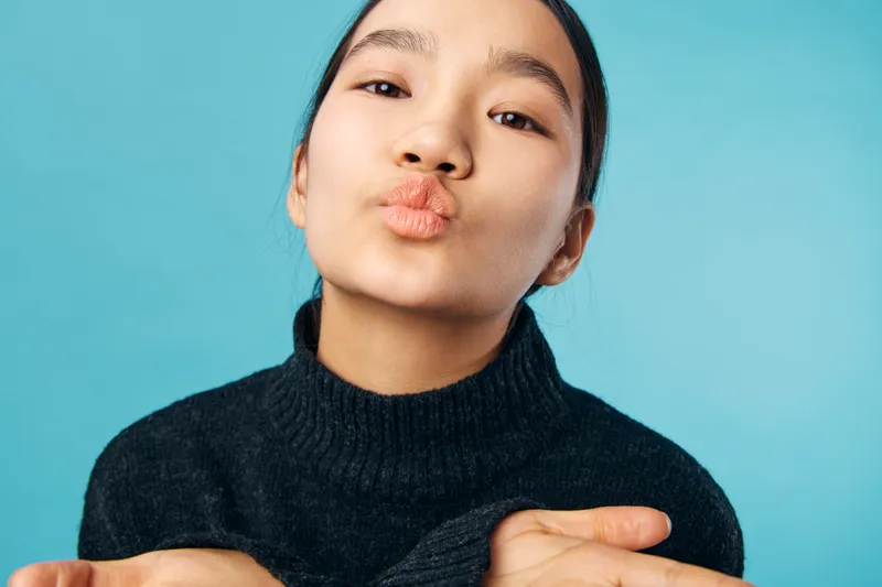 Young woman with black sweater blowing kiss and looking at camera against blue background. Closeup portrait of female person sending love or affection with puckered lips and casual style.