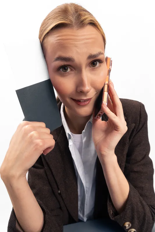 Young woman in business suit talking on smartphone, holding black notebook, smiling with confident expression. Concept for business communication, multitasking, and professional lifestyle.