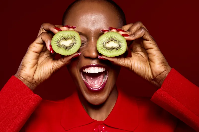 Happy African woman with dark skin holds two kiwi halves in front of her eyes, smiling widely. Vibrant red clothes and background create joyful and fresh concept of healthy food and wellness.