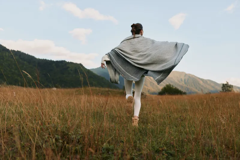 Woman in flowing cape walks across a grassy field mountains in the distance capturing an outdoor lifestyle moment of freedom movement and connection with nature today