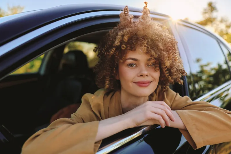 A smiling woman leans from a car window during golden hour, captured with retro film color. A candid lifestyle portrait conveying freedom, relaxation, and everyday travel vibes.