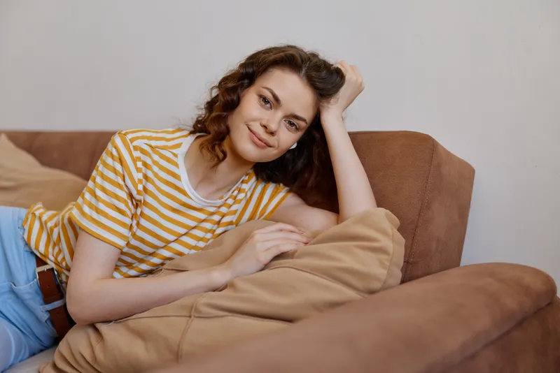 Calm woman relaxing on a sofa at home, wearing a striped yellow T shirt, soft cushions nearby, showcasing a cozy, casual lifestyle scene in a bright living room with natural light