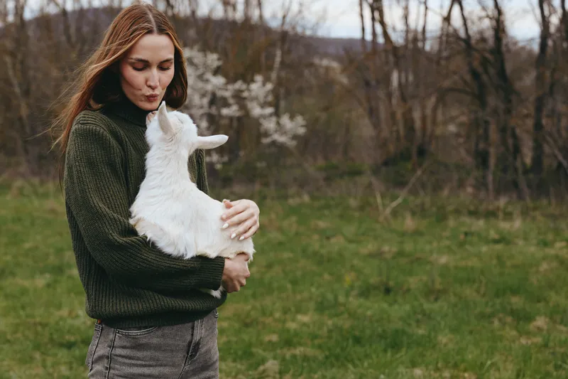 Goat kid held by a woman outdoors, tender moment in a grassy field, cozy sweater, gentle smile, soft natural light, rustic backdrop, nature and connection inspiring calm lifestyle scene