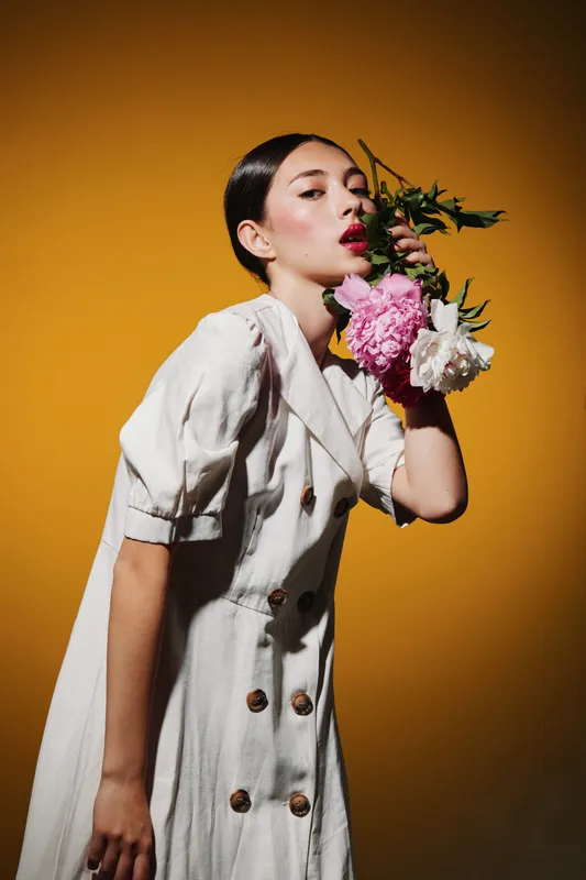Young woman in a stylish cream dress holds pink and white flowers near face against a bold yellow backdrop. Editorial fashion concept, creative beauty portrait with modern styling.