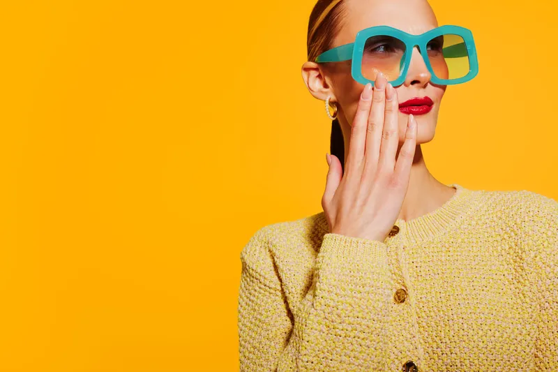 Stylish woman in yellow sweater and large turquoise glasses touches her face against bright yellow background. Concept of fashion, beauty, accessories, and confident modern style.