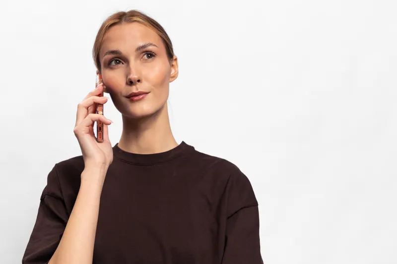 Young woman in brown shirt talking on smartphone, looking thoughtful on white background. Concept of communication, technology, and casual lifestyle.