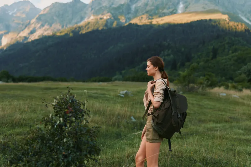Woman hiker with backpack stands in a meadow with distant mountains, capturing a lifestyle moment in nature, calm pose, adventurous spirit, solo exploration, outdoor