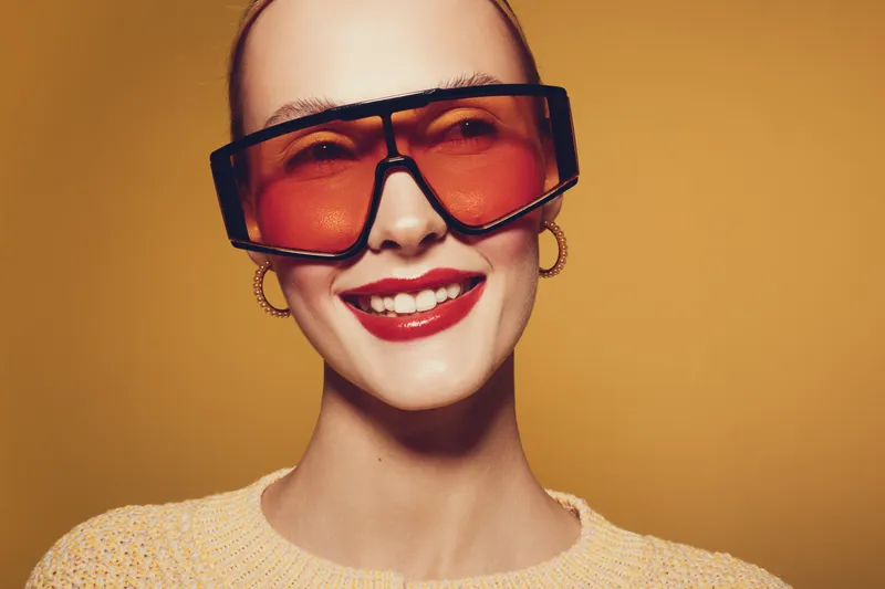 Smiling woman with large orange sunglasses and red lipstick poses against yellow background. Fashion portrait with trendy eyewear and joyful expression in studio lighting.