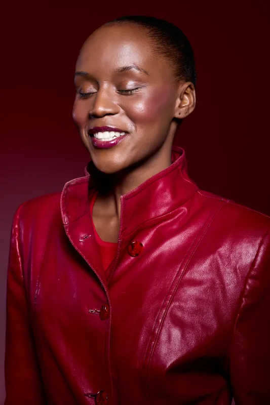 Portrait of a smiling African woman with dark skin wearing a red leather jacket on a red background. Concept of fashion, beauty, confidence, and elegance in studio lighting.