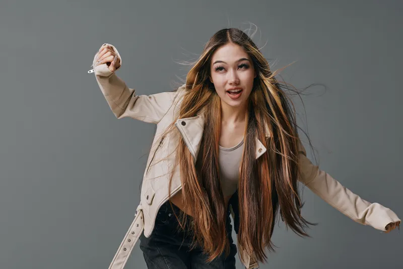 Young woman with long hair wearing light jacket and casual outfit poses dynamically against gray background. Female model with expressive face and movement in studio lighting.