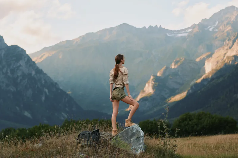 A woman stands on a rock in a mountain meadow, ready for a hike. She wears casual outdoor attire and gazes toward rugged peaks, embodying solo travel and nature immersion.