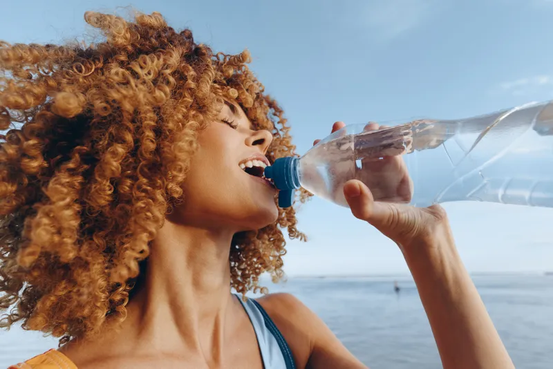 Woman with curly hair drinking water from a clear bottle, close up outdoors near the sea, bright summer sunlight, refreshing hydration concept, wellness and self care, joyful smile, today